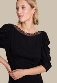 Svart kjole med puffermer og båtutringning med blonder. Har dekorativ blonde i midjen. Mykt stoff og tettsittende fasong.