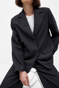Blazer noir à rayures avec un design à un bouton, poches avant et fermeture à bouton. Fabriqué à partir d'un tissu texturé, associé à un haut blanc.