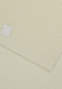 Écharpe en tricot de couleur crème avec un motif texturé, comportant une petite étiquette en tissu blanc avec un emblème décoratif dans un coin.