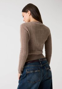 Brauner gerippter Strickpullover mit figurbetontem Schnitt, hohem Ausschnitt und langen Ärmeln, kombiniert mit dunkelblauen Jeans mit Reißverschlusstaschen auf der Rückseite.