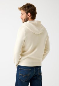 Sudadera con capucha crema y ribete acanalado, combinada con jeans azul oscuro que tienen bolsillos traseros y costuras amarillas. Tejido suave y texturizado.