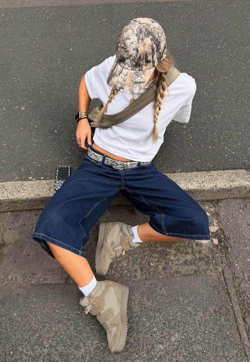 Personne aux cheveux tressés portant une casquette camouflage, une chemise blanche, un short en denim ample et des baskets hautes beige, assise sur un trottoir.