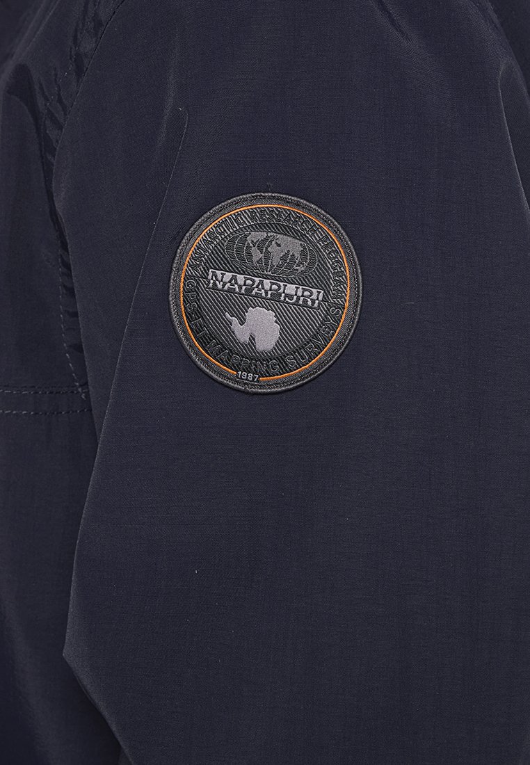 Manga de chaqueta azul marino oscuro con un parche circular negro y naranja que presenta un motivo del mundo y el texto "NAPAPIJRI 1987". Textura de tela suave.