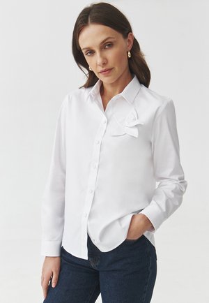 Camicia bianca con bottoni realizzata in tessuto liscio, dotata di un accento a fiocco sul lato sinistro del petto, con maniche lunghe e colletto classico.