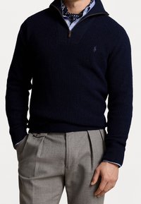 Mörkblå stickad tröja med kvartdragkedja, strukturerat mönster och en liten logotyp på bröstet, i kombination med ljusgrå plisserade byxor.