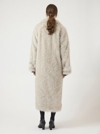 Γυναίκα με καφέ αλογοουρά, φορώντας ένα μακρύ μπεζ φουσκωτό παλτό, μαύρες καλσόν και μαύρες ψηλές γόβες, στέκεται με την πλάτη στραμμένη προς την κάμερα.