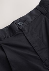 Juodos medžiagos kelnės su lygia priekyje, subtiliomis klostėmis ir sagos užsegimu liemenyje, pasižyminčios lygiu paviršiumi.