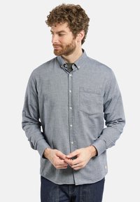 Homme aux cheveux bouclés et à la barbe portant une chemise grise à manches longues boutonnée et un jean noir, regardant vers sa gauche avec les mains jointes.