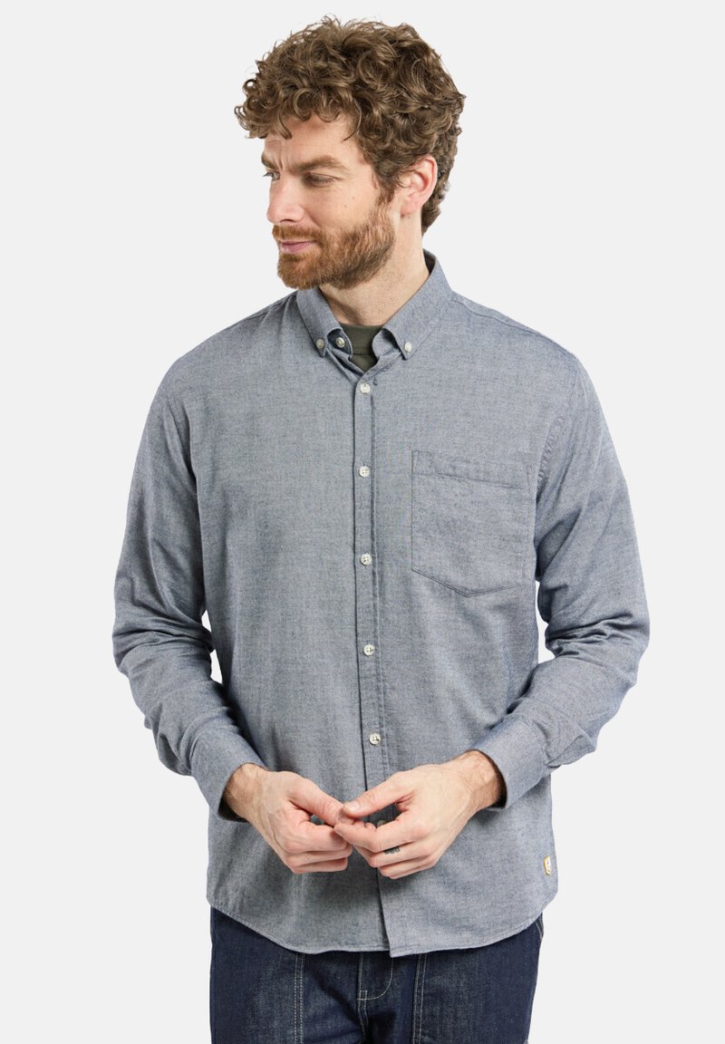 Homme aux cheveux bouclés et à la barbe portant une chemise grise à manches longues boutonnée et un jean noir, regardant vers sa gauche avec les mains jointes.