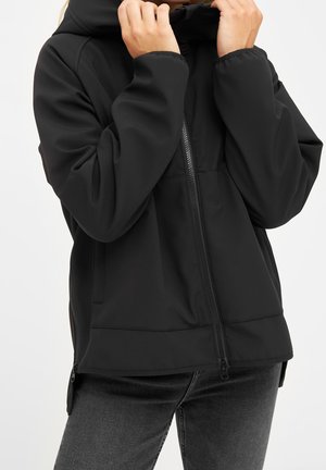 Personne portant une veste noire zippée avec capuche, les mains ajustant le col, associée à un jean gris foncé, sur fond blanc.