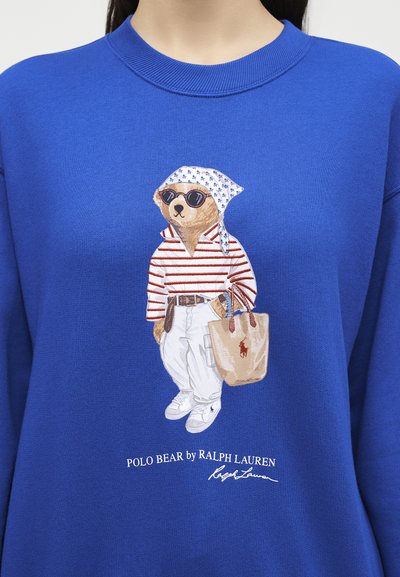 Αρκούδα με γυαλιά ηλίου, ριγέ πουκάμισο, λευκό παντελόνι, αθλητικά παπούτσια, μαντήλι στο κεφάλι, κρατώντας μπεζ τσάντα, τυπωμένη σε μπλε φούτερ με κείμενο Polo Bear by Ralph Lauren.