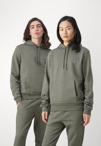 Sudaderas con capucha en verde oliva de tela suave, con un bolsillo frontal y un pequeño logotipo bordado en el pecho, combinadas con pantalones a juego.