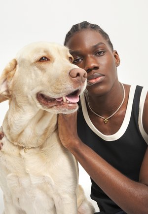 Persona joven con cabello trenzado que lleva una camiseta sin mangas negra, sosteniendo suavemente a un gran Labrador amarillo, ambos mirando hacia adelante sobre un fondo blanco.