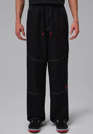 Man draagt zwarte losse broek met witte stiksels, trekkoord in de taille en een rood logo bij de linkerenkel, gecombineerd met zwarte schoenen met rode zolen.