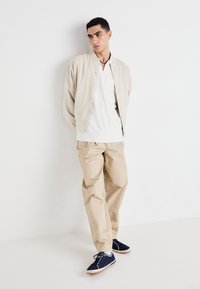 Lys beige jakke med knapknapper, hvit ribbet undertrøye, beige løstsittende bukser og marineblå sneakers med hvite detaljer.