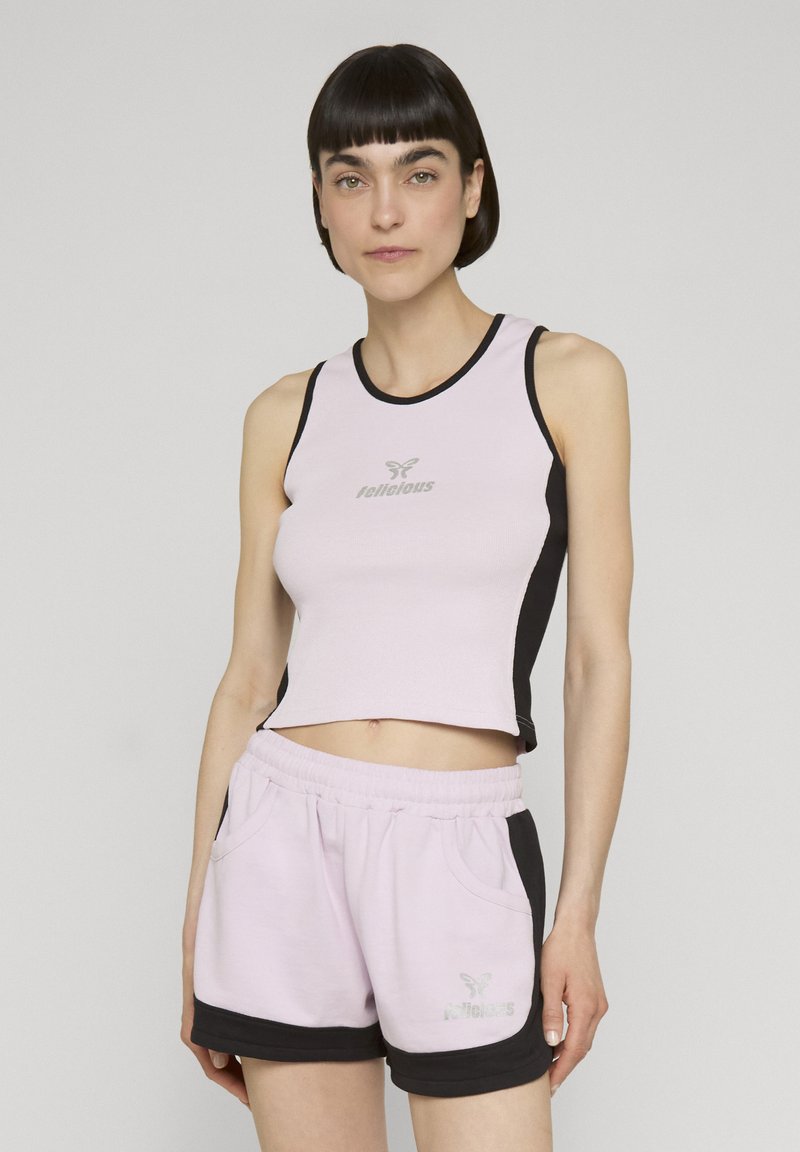 Femme aux cheveux courts et foncés portant un crop top rose et noir sans manches et un short assorti avec les logos de la marque sur les deux vêtements.
