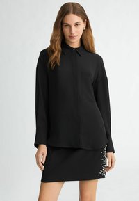 Black long-sleeve blouse with a collar, boxy fit, and single chest pocket, paired with a fitted black skirt featuring pearl accents on one side.