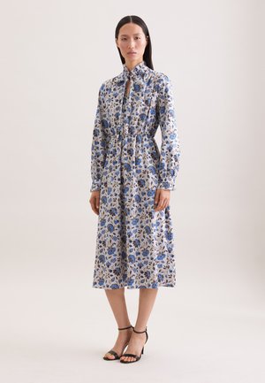 Kobieta w niebieskiej, kwiatowej sukience midi z długim rękawem i wiązanym dekoltem, stojąca na jasnym, jednolitym tle w czarnych sandałach na obcasie.