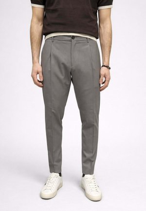 Uomo con pantaloni grigi aderenti, calzini neri, scarpe da ginnastica bianche, camicia marrone scuro e braccialetto nero, in piedi davanti a uno sfondo semplice.