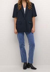 Blazer de gran tamaño en color marino con mangas cortas, cierre de un solo botón y dos bolsillos frontales, combinado con jeans rectos de color azul claro y botas negras hasta el tobillo.