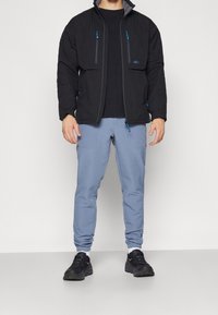 Veste rembourrée noire avec des accents gris et des zips bleus, associée à un pantalon bleu clair avec des poches latérales et des poignets élastiques, portée avec des chaussures noires.