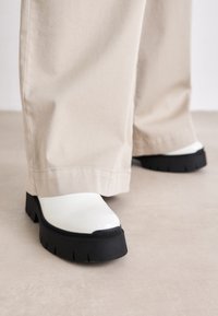 Pantofi albi impermeabili cu o talpă grosolană din cauciuc negru, având un finisaj texturat și un detaliu logo, asortați cu pantaloni largi bej.