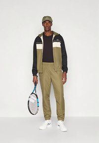 Fato de treino desportivo em verde-oliva e preto, com casaco de fecho éclair com riscas brancas e pretas e calças cónicas combinando; segurando uma raquete de ténis.