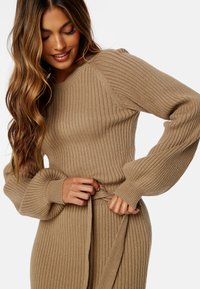 Maglione a coste in tonalità beige con maniche lunghe a sbuffo e cintura in vita. Presenta un tessuto texturizzato e un design aderente.
