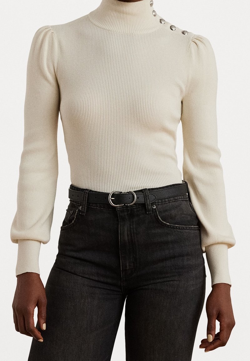 Pull à col roulé côtelé beige avec épaules bouffantes et boutons décoratifs, associé à un jean noir taille haute et une ceinture noire.