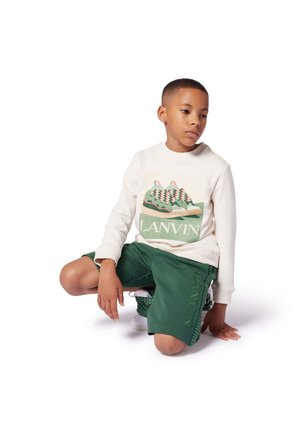Felpa bianca con grafica di sneaker e testo "LANVIN"; pantaloni corti verdi con dettaglio logo; ragazzo giovane in posa su uno sfondo bianco.