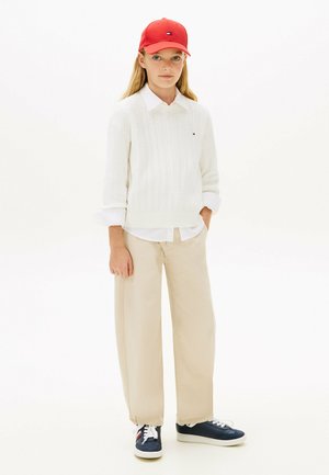 Jong persoon met een rode pet, witte gebreide trui over een wit shirt, beige wijde broek en donkerblauwe sneakers, staand met één hand in de zak.