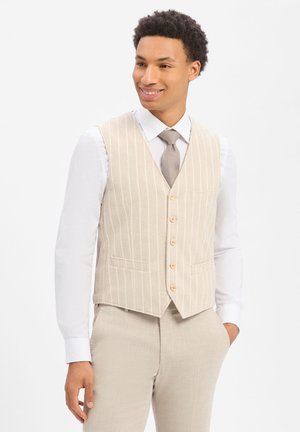 Mann trägt eine beige Nadelstreifenweste und passende Hose, weißes Hemd und hellgraue Krawatte, lächelt und schaut seitlich.