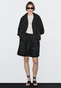 Sorte letvægtsjakke med bred krave, grå skjorte, sorte plisserede shorts og sorte stropsandaler. Minimalistisk design, glatte teksturer.