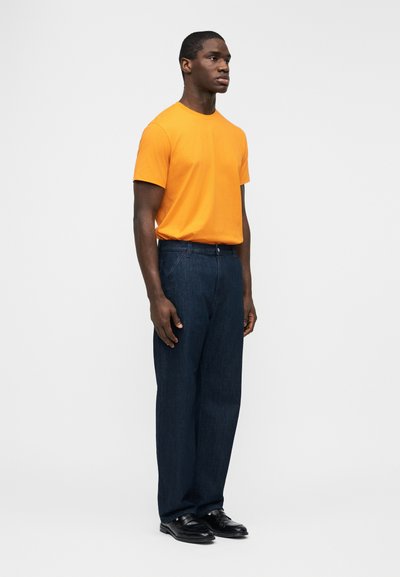 Άνδρας όρθιος με πορτοκαλί κοντομάνικη μπλούζα, σκούρο μπλε τζιν και μαύρα παπούτσια μπροστά σε απλό ανοιχτό γκρι φόντο.