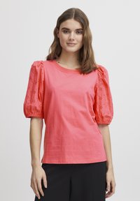 Tricou din bumbac corai cu mâneci scurte bufante, având detalii cu găurele, guler rotund și croială lejeră. Potrivit cu pantaloni negri.