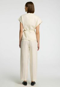 Lichtbeige linnen top met korte mouwen, aangetrokken taille en brede beige linnen broek. Model draagt zwarte schoenen; effen achtergrond.