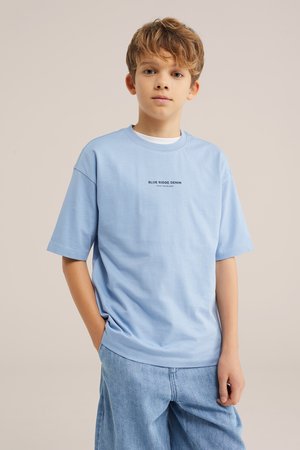 Poiss helepruuni juustega, kandes helesinist laiema vabaaja T-särki ja lõdvaid teksapükse, seisab ühevärvilise beeži tausta ees.