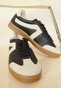Must mustad ja kreemjad sneakerid, mis on tehtud nahast ja kummist, omavad tekstuuriga talla, ümarat varvast ja musti shoelace'e, millel on kontrastsed disainielemendid.
