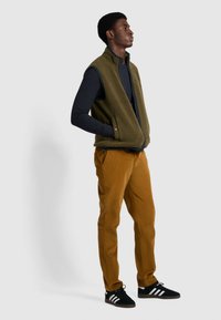 Grüne Fleece-Weste mit Reißverschluss, navyblaues Langarmshirt, beige Cordhosen und schwarze Sneaker mit weißen Streifen. Stehende Pose, neutraler Hintergrund.