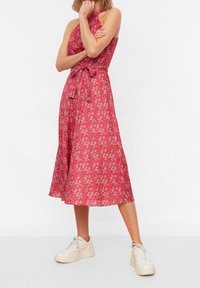 Kvinna som bär en röd ärmlös midiklänning med små vita blommönster och vita plattformssneakers, stående med ena handen på hakan.