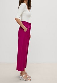 Fuksjowe spodnie o szerokich nogawkach z plisami z przodu i bocznymi kieszeniami, zestawione z dopasowanym białym topem z prążkowanego materiału oraz białymi sandałami na obcasie z paskami.