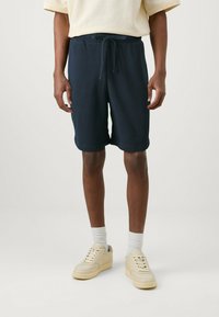 Shorts in cotone misto blu navy con vita elasticizzata e cordino. Tessuto liscio, taglio a ginocchio, abbinati a sneakers crema e calzini bianchi.
