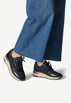 Sneakers noirs et bleu marine avec une tige texturée, fermeture à lacets et semelle beige. Des accents détaillés mettent en valeur un design sportif. Associés à un pantalon en denim large.
