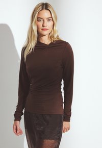 Moteris su ilgais blondiniais plaukais, vilkinti prigludusią rudą ilgomis rankovėmis palaidinę ir permatomą tamsiai rudą sijoną prieš baltą foną.