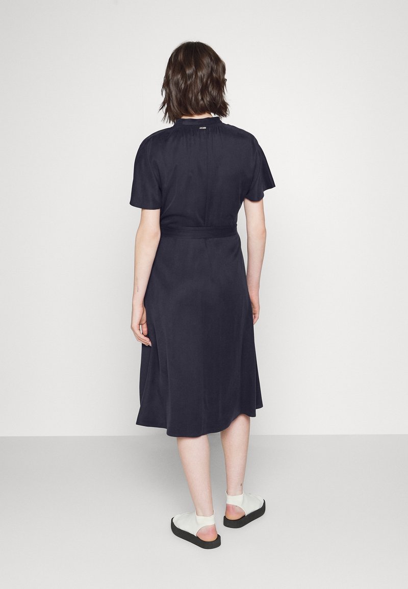 Personne aux cheveux courts et foncés portant une robe bleu marine longueur genou et des chaussures blanches ouvertes à l'arrière, debout face à un mur blanc uni.