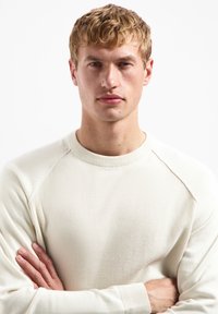 Crème-kleurige trui met een gestructureerd breipatroon, geribbelde kraag en raglanmouwen; model poseert met gekruiste armen voor een witte achtergrond.
