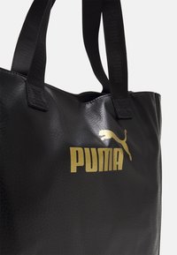 Sac fourre-tout noir avec surface texturée, poignées en tissu noir et logo Puma doré imprimé sur le devant.