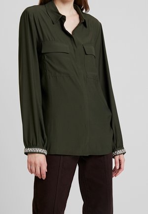 Femme portant une chemise boutonnée à manches longues vert foncé avec des poches poitrine et des poignets décoratifs à motifs noir et blanc, assortie à un pantalon foncé.