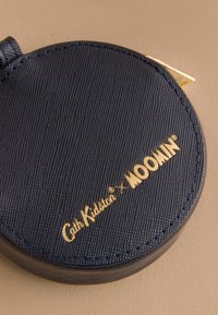 Kulatá kožená peněženka na mince navy barvy s texturovaným povrchem, zdobená zlatým embosovaným logem "Cath Kidston x MOOMIN" a kovovým zipem.