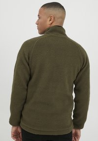 Fleece jacket in olive green, featuring a high collar and textured surface. The fit is relaxed, with long sleeves and a rounded hemline.